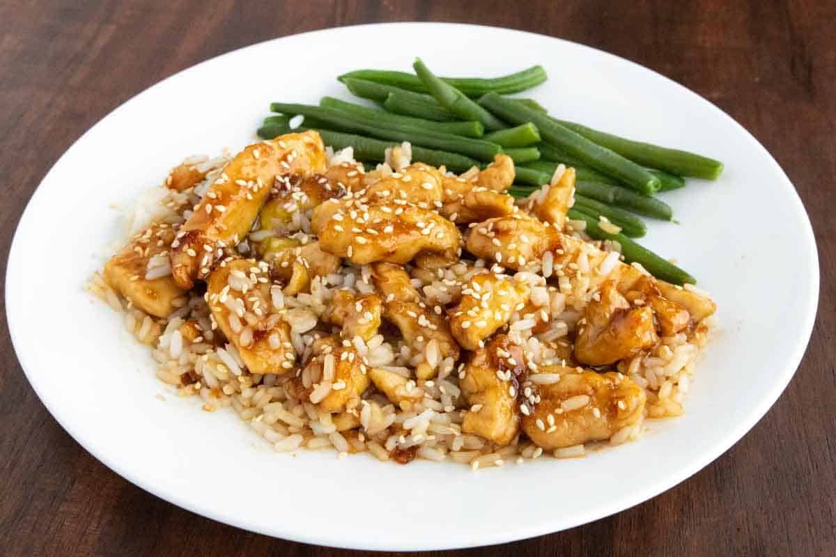 Bourbon chicken on a bed of rice on a white plate with green beans.