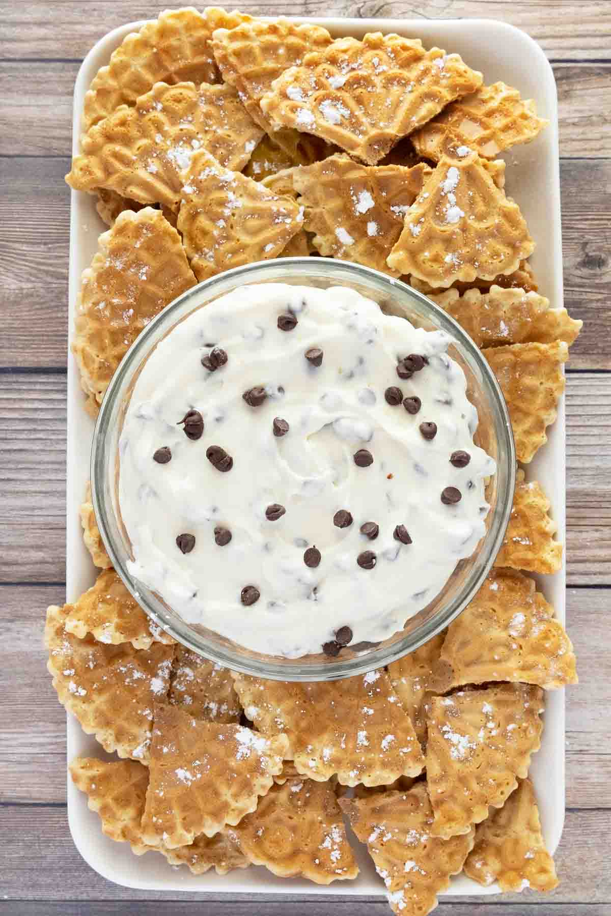 Cannoli dip in a glass bowl with pizzelle quarters on a white serving platter.