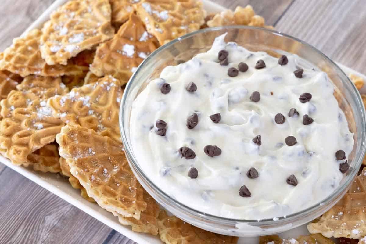 Cannoli dip in a glass bowl with pizzelle quarters on a white serving platter.