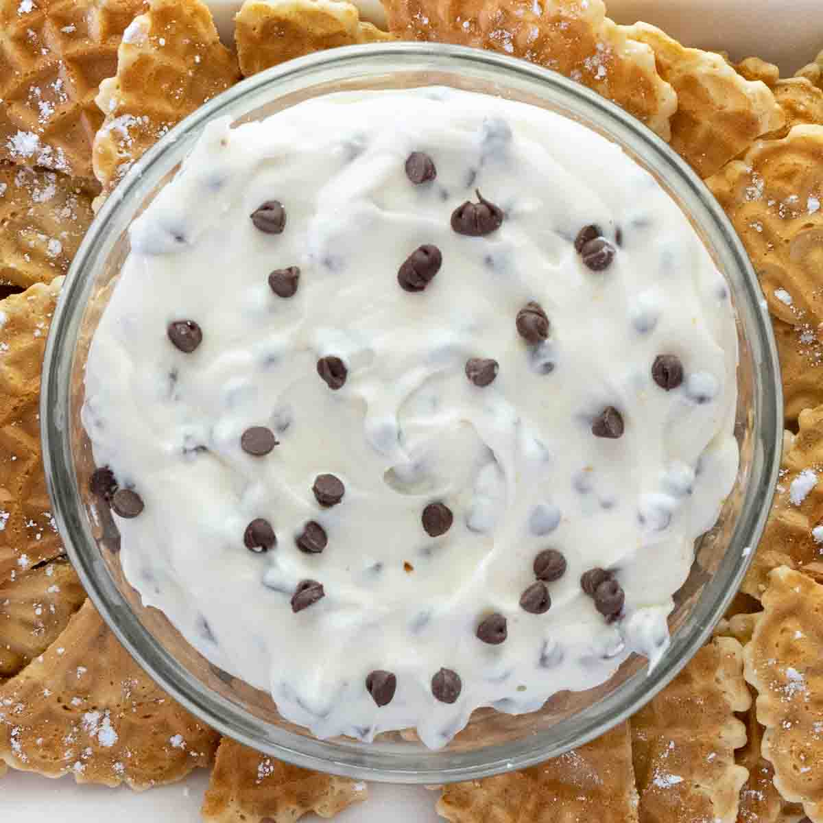 Cannoli dip in a glass bowl with pizzelle quarters on a white serving platter.