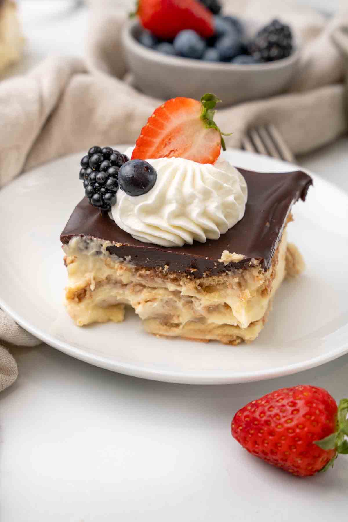 Slice of eclair cake on a white plate with berries in the foreground and background.