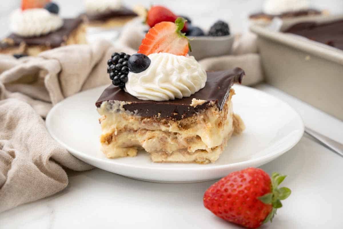 Slice of eclair cake on a white plate with berries in the foreground and background.