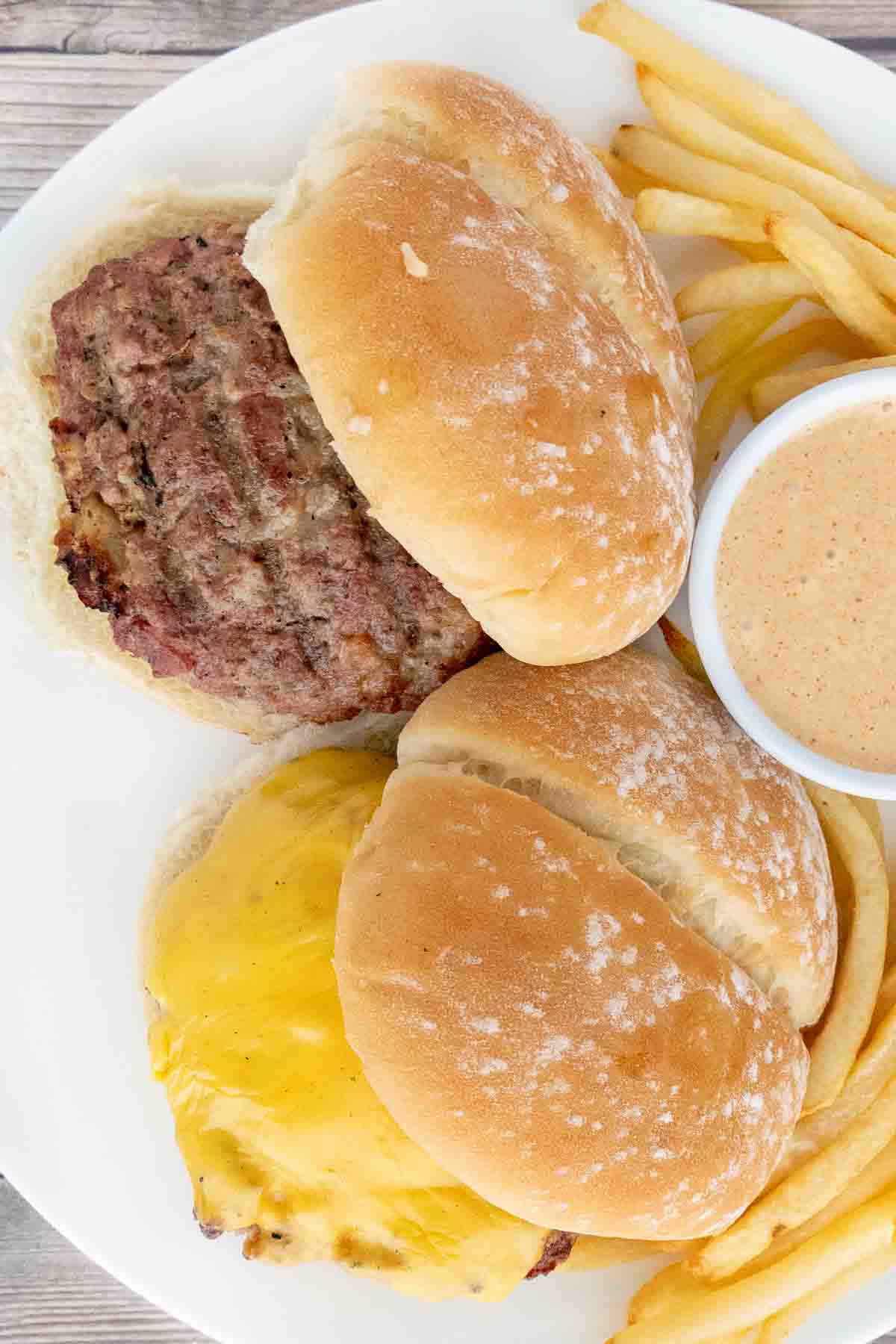 One crack burger and one with cheese on buns on a white plate with french fries and sauce.