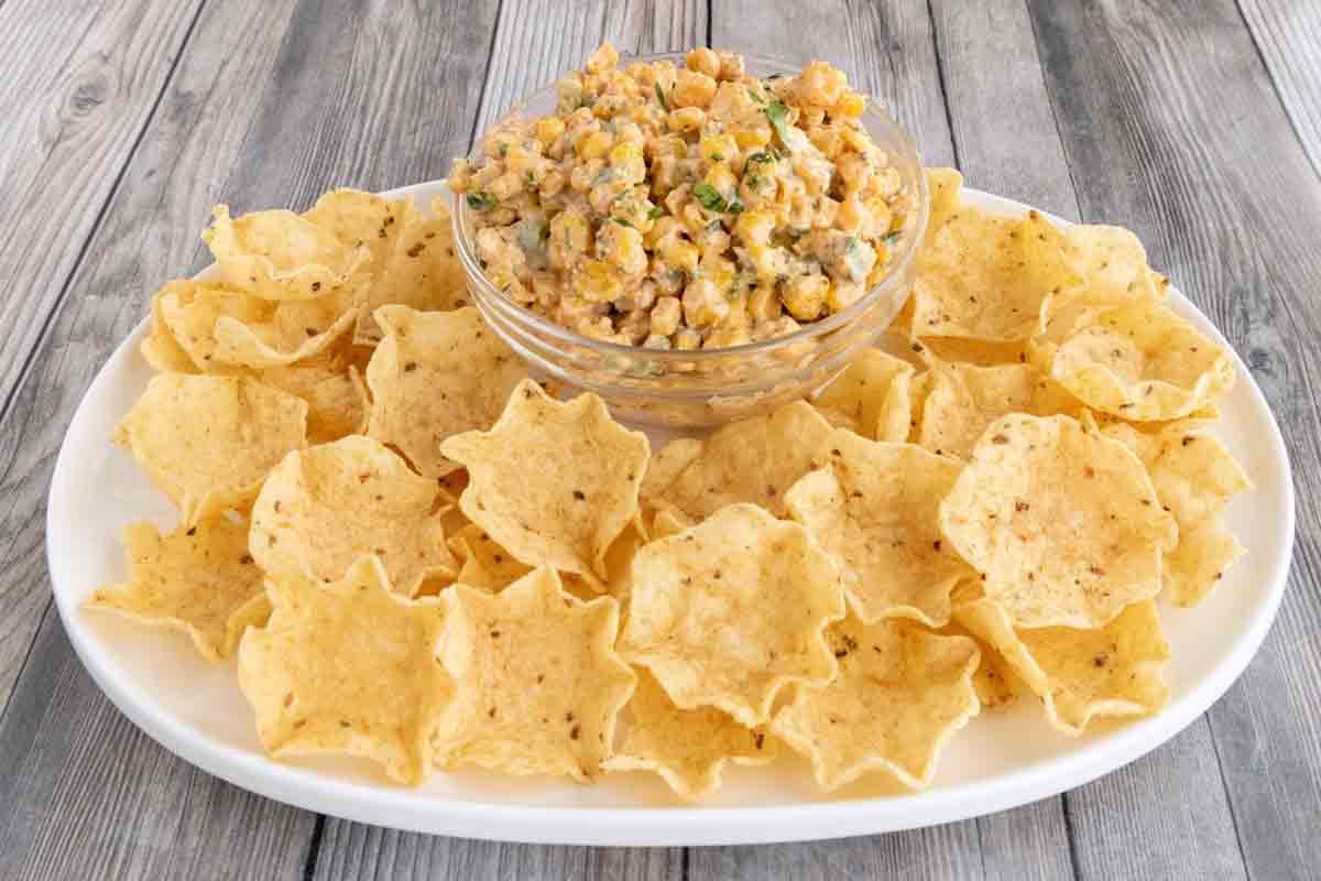 Elotes dip in a glass bowl with tortilla chips on a white platter.