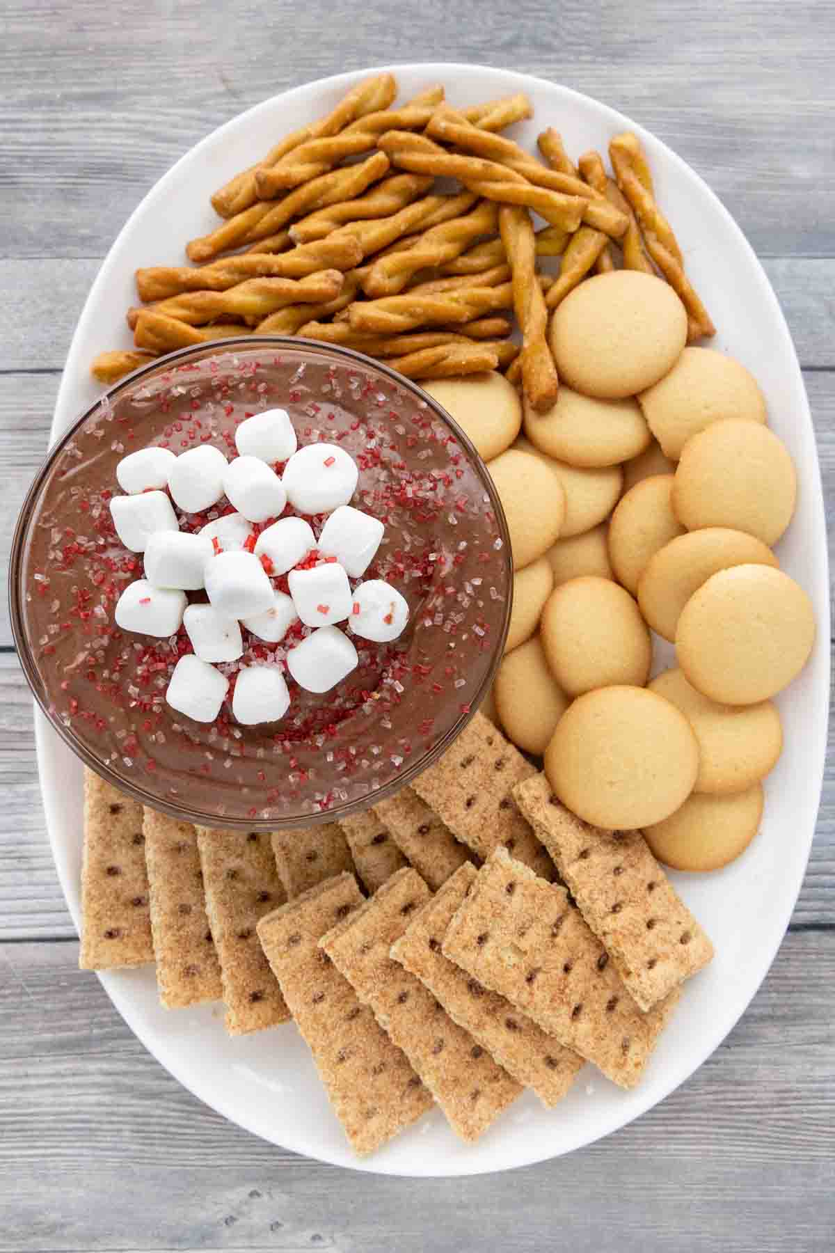 Hot cocoa dip with cookies and pretzels on a white plate.