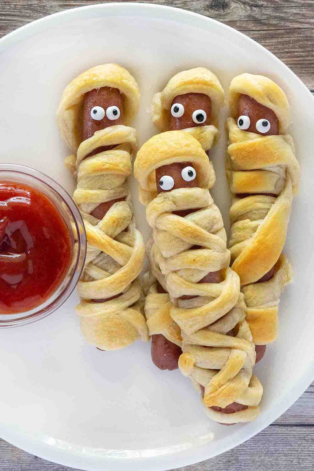 Mummy dogs on a white plate with a bowl of ketchup.