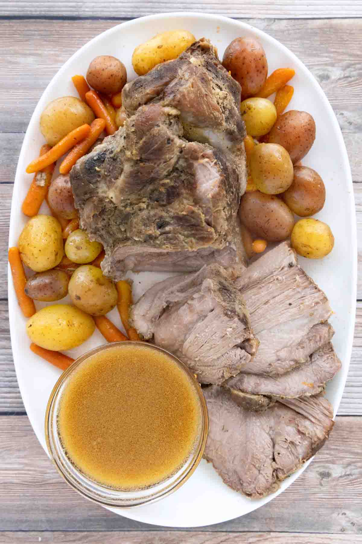 Slow cooker pork loin sliced on a white platter with potatoes, carrots and au jus.